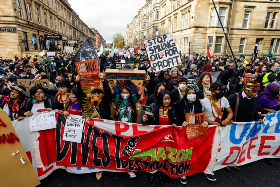 Manifestação do Dia de Ação Global em Glasgow, na Escócia. Na frente, vemos lideranças indígenas do Brasil com cartazes coloridos que chamam a atenção para as ações contra a mudança climática.